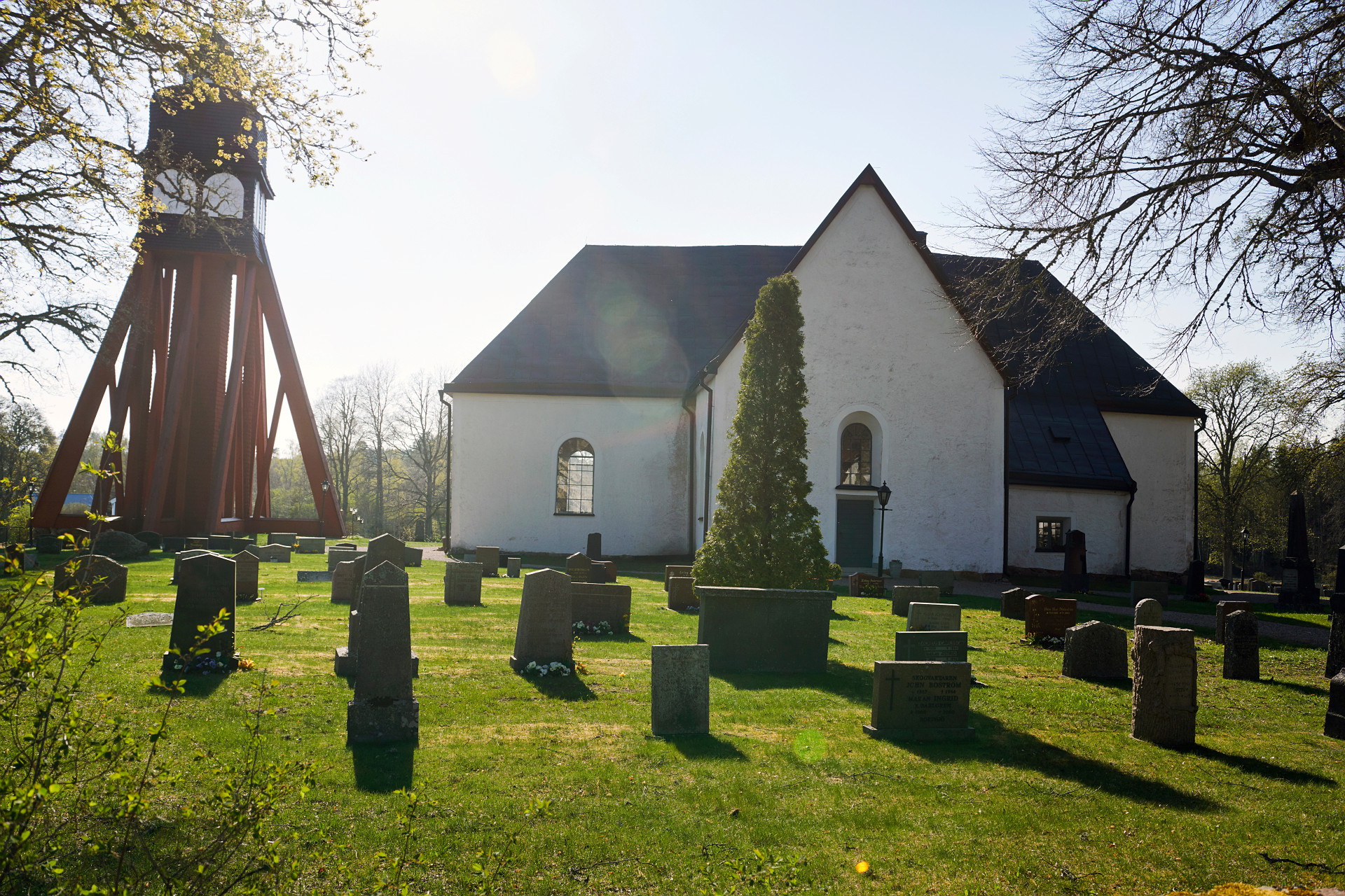 Solen skiner ner på ett klocktorn i rött och en vit ganska kompakt kyrka. I förgrunden ser vi kyrkogården med gravstenar och grönområden.