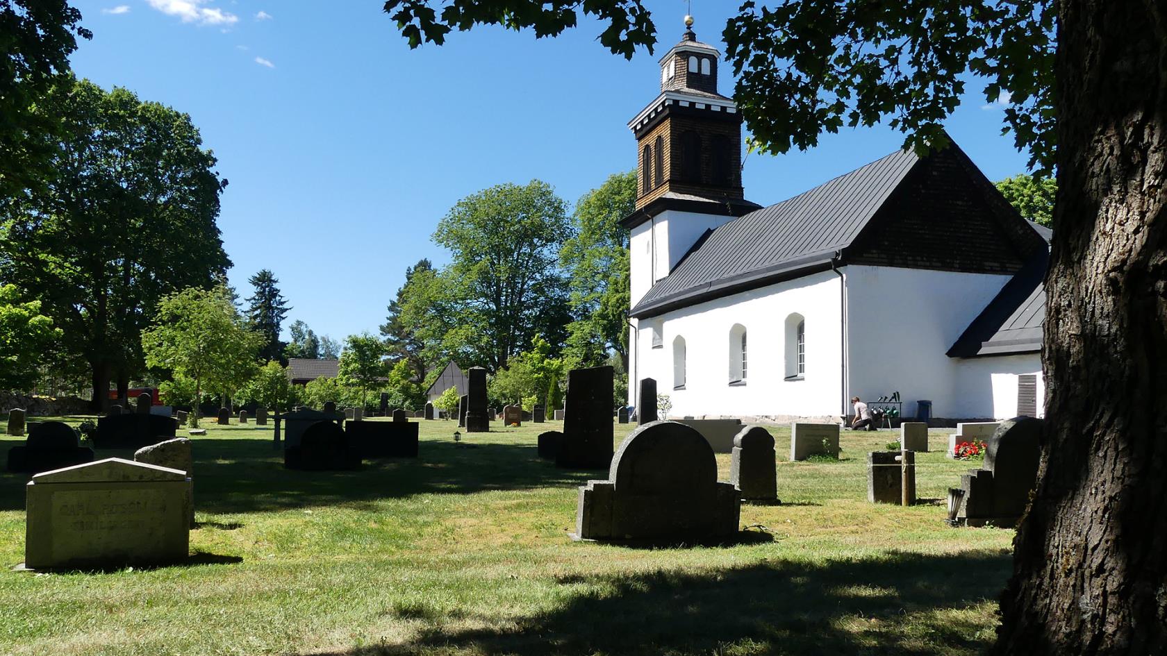 I bakgrunden en vit stenkyrka med svar tak. Sen ser vi delar av kyrkogården och i förgrunden ett träd och dess skugga.