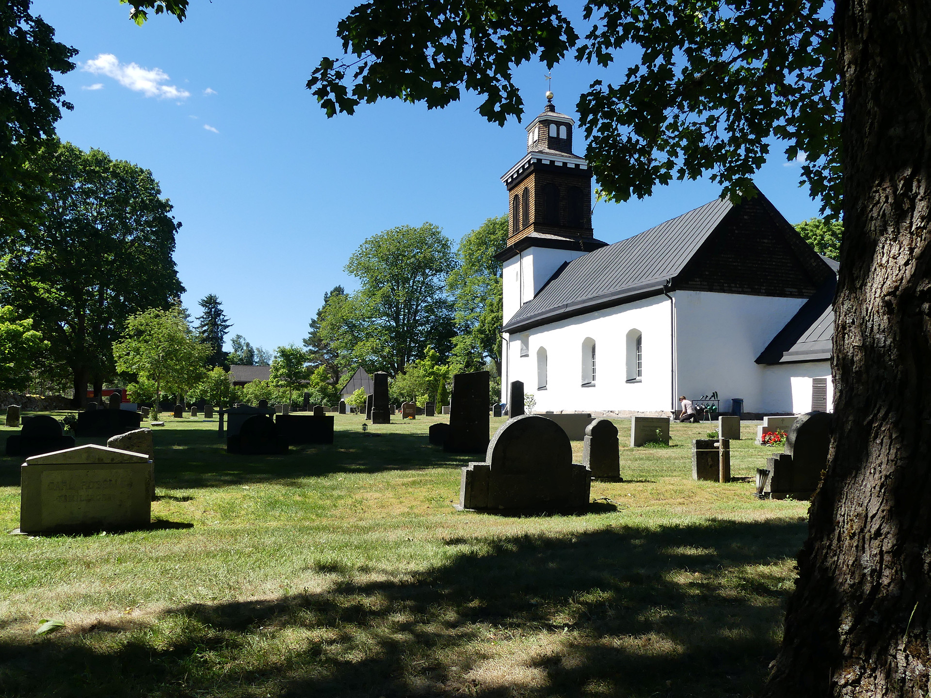 I bakgrunden en vit stenkyrka med svar tak. Sen ser vi delar av kyrkogården och i förgrunden ett träd och dess skugga.