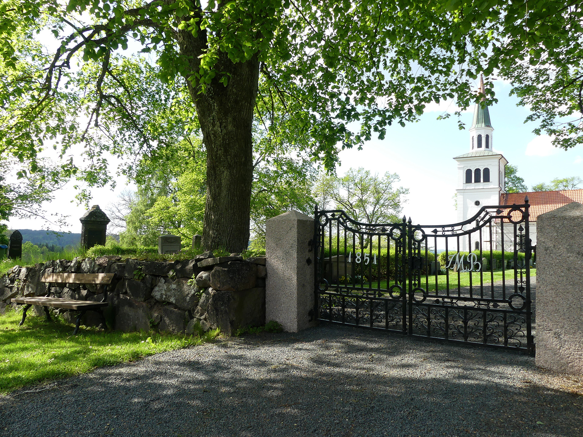 En kyrkmur med en bänk framför till vänster i bild. De stängda grinden är i svart smide. Mitt i bild bakom muren mellan bänken och grinden ett grönskande träd. I bakgrunden skymtar vi en vit stenkyrka. 