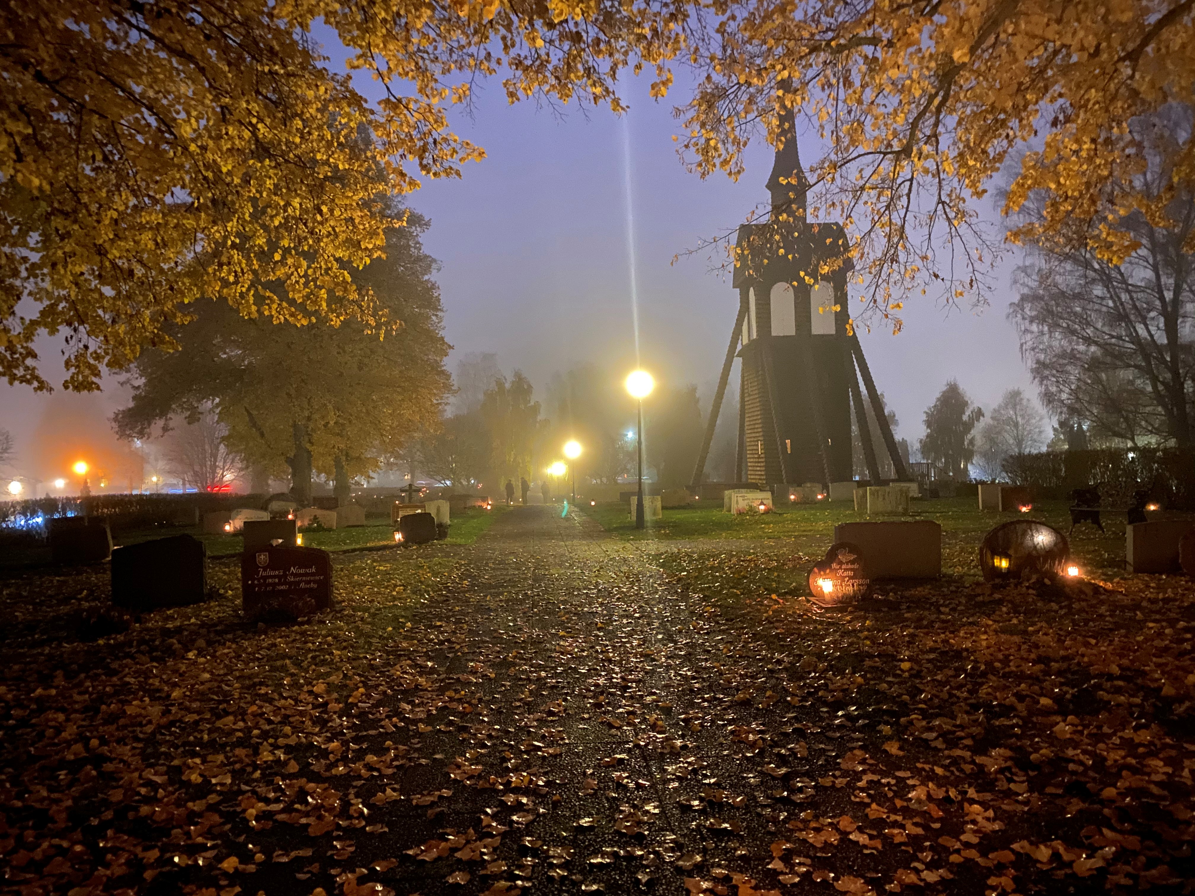 Bild på en kyrkogård med klocktornet uppe i vänstra hörnet. Det är allhelgonstad - mörkt med en massa tända ljus på gravarna.