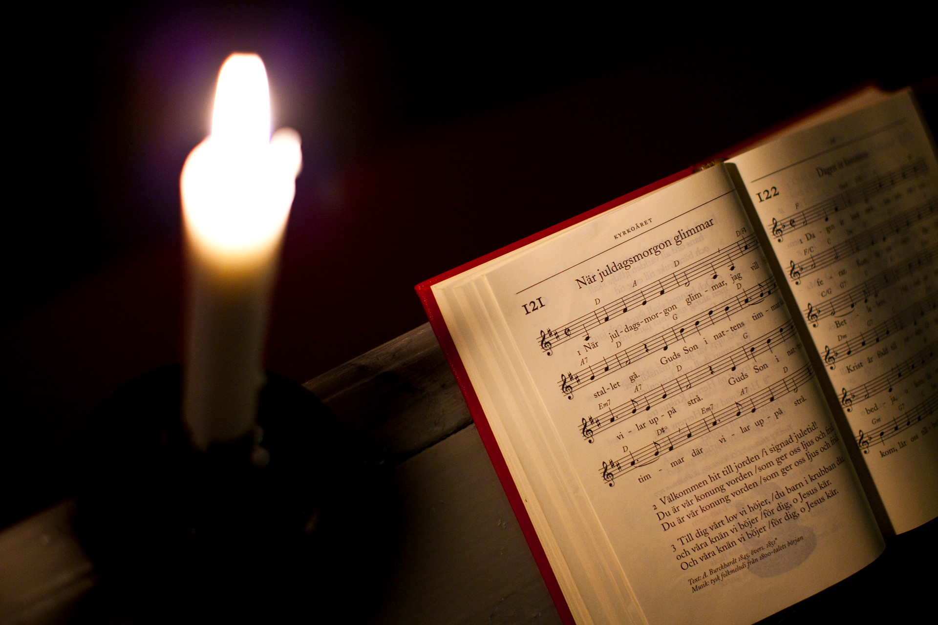 Ett levande ljus och en psalmbok i en mörk kyrka. Psalmen är "När juldagsmorgon glimmar".