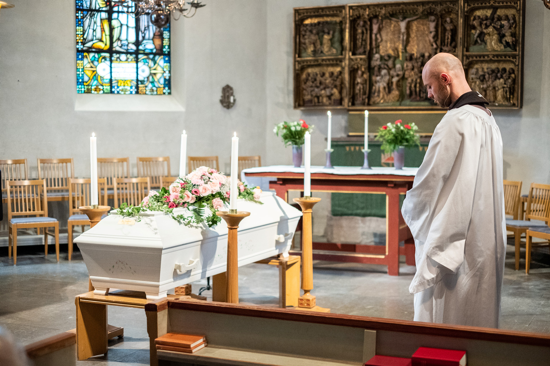 En kyrksal med en vit kista med rosa rosor på locket står till vänster i bild. På andra sidan om kistan står en präst i liturgiska kläder och med nerböjt huvud. I förgrunden syns delar av kyrkbänkar i trä.