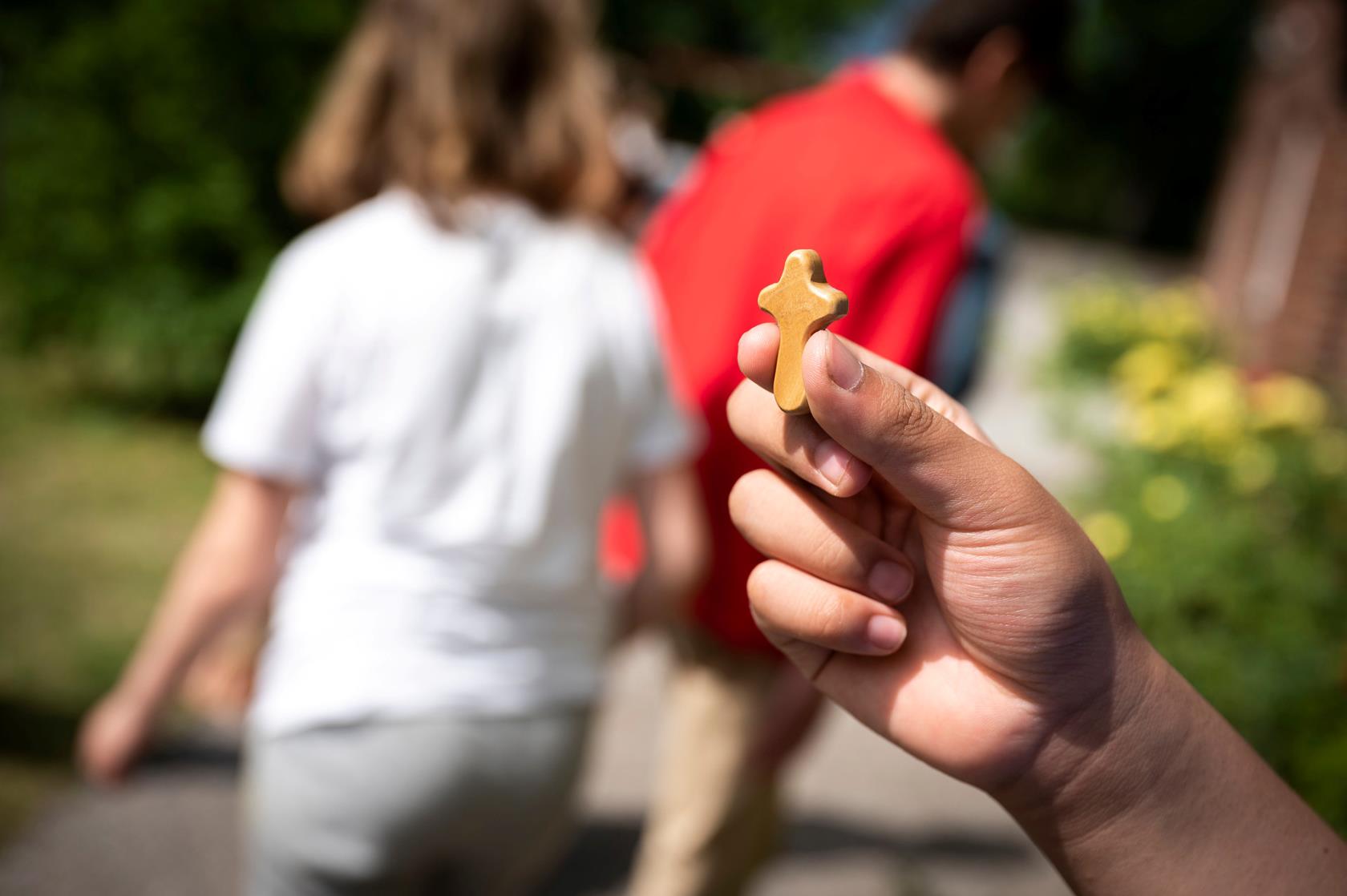 En hand som håller i ett litet träkors. I bakgrunden två personer som går iväg.