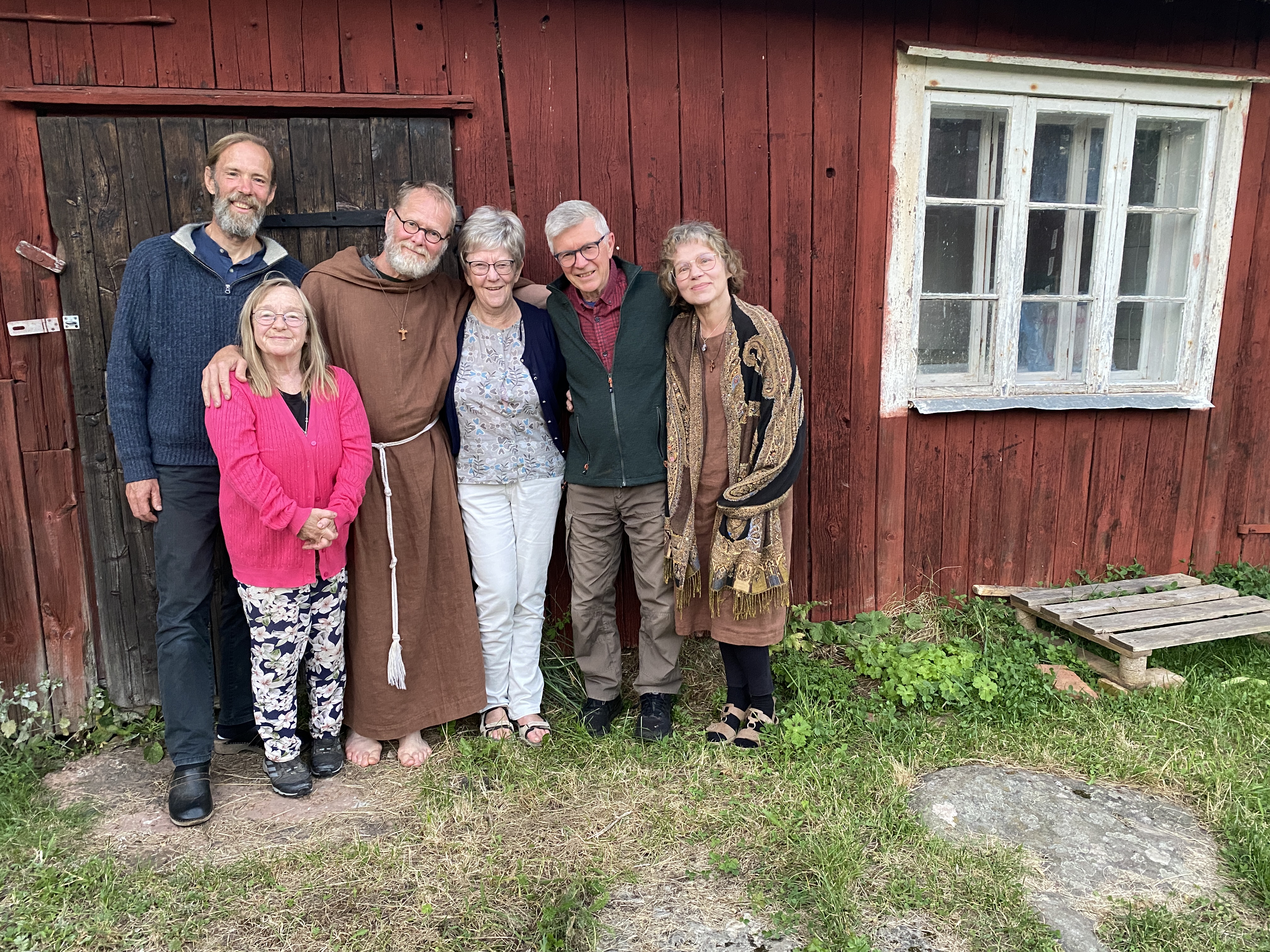 En grupp människor i en rad, de ler och håller om varandra. Bakom dem är ett rödmålat hus.