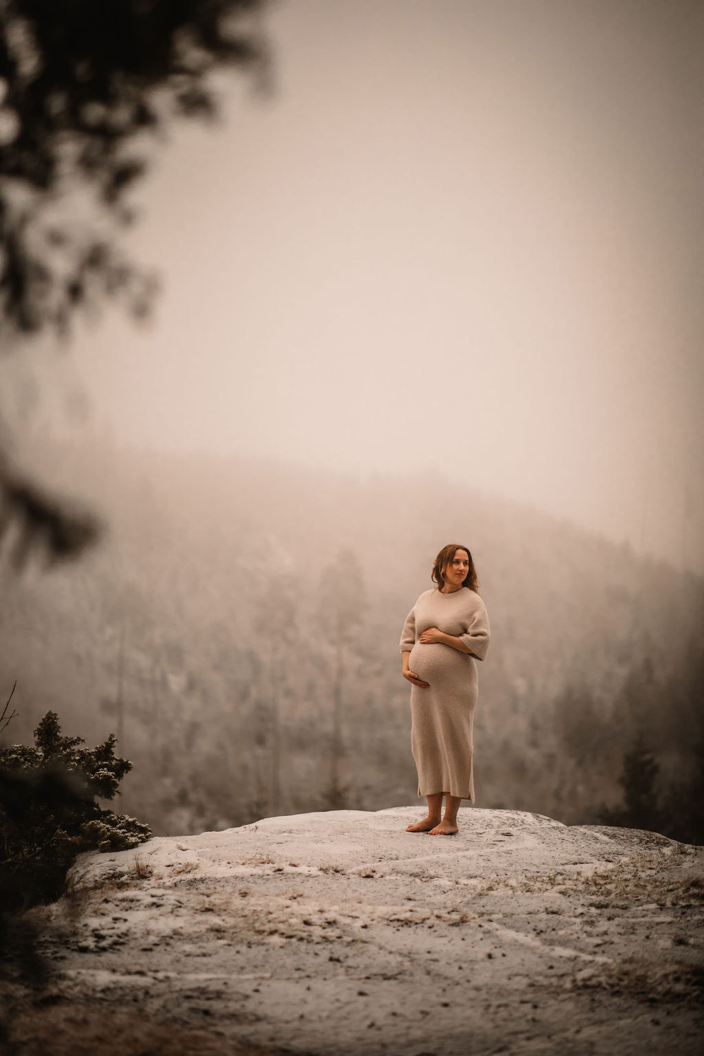 En gravid kvinna står barfota på en klippa och håller händerna på magen. 