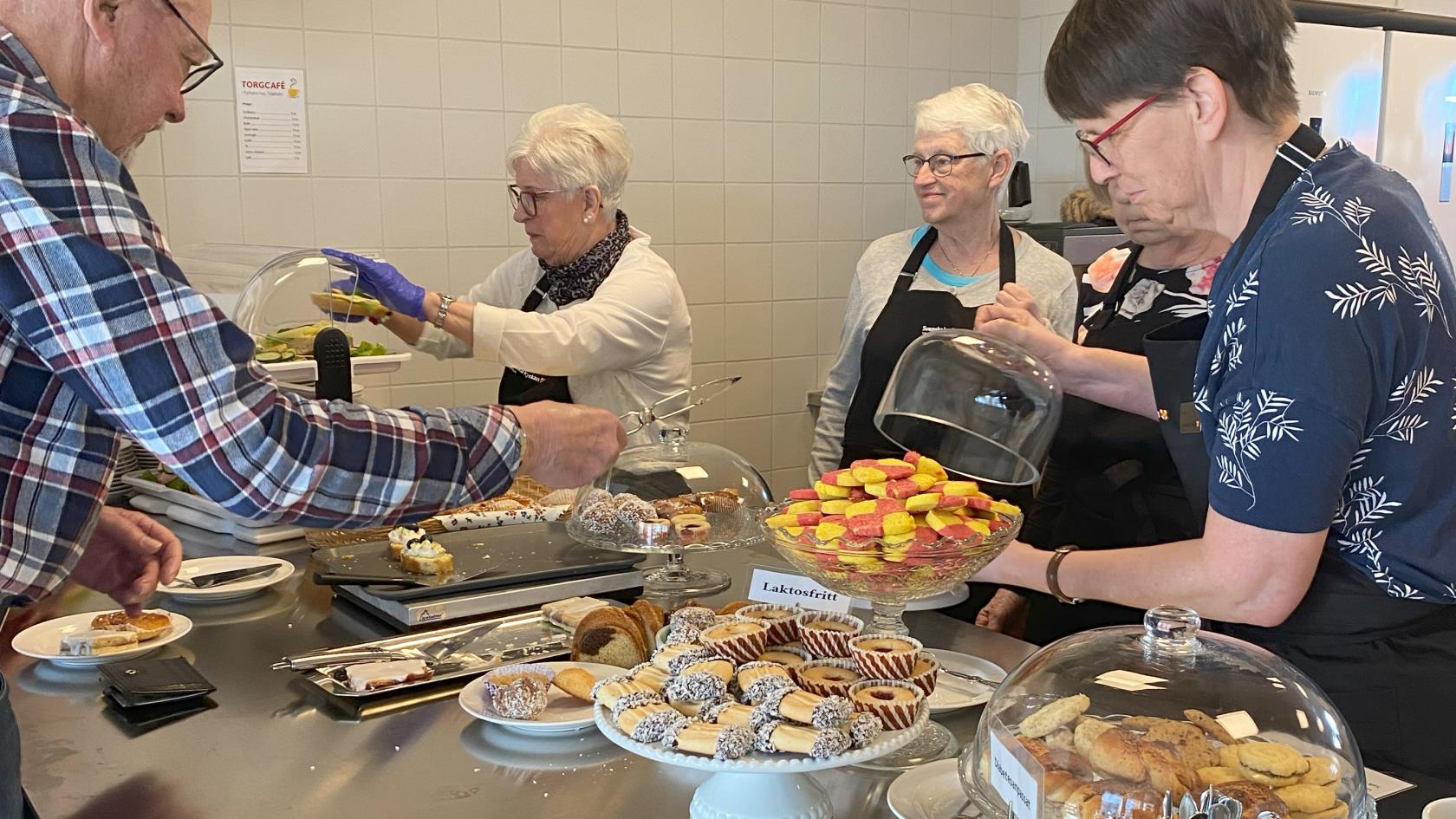 Volontärer i svarta förkläden med Svenska kyrkan Tidaholms logga på hjälper torgcafékunder.