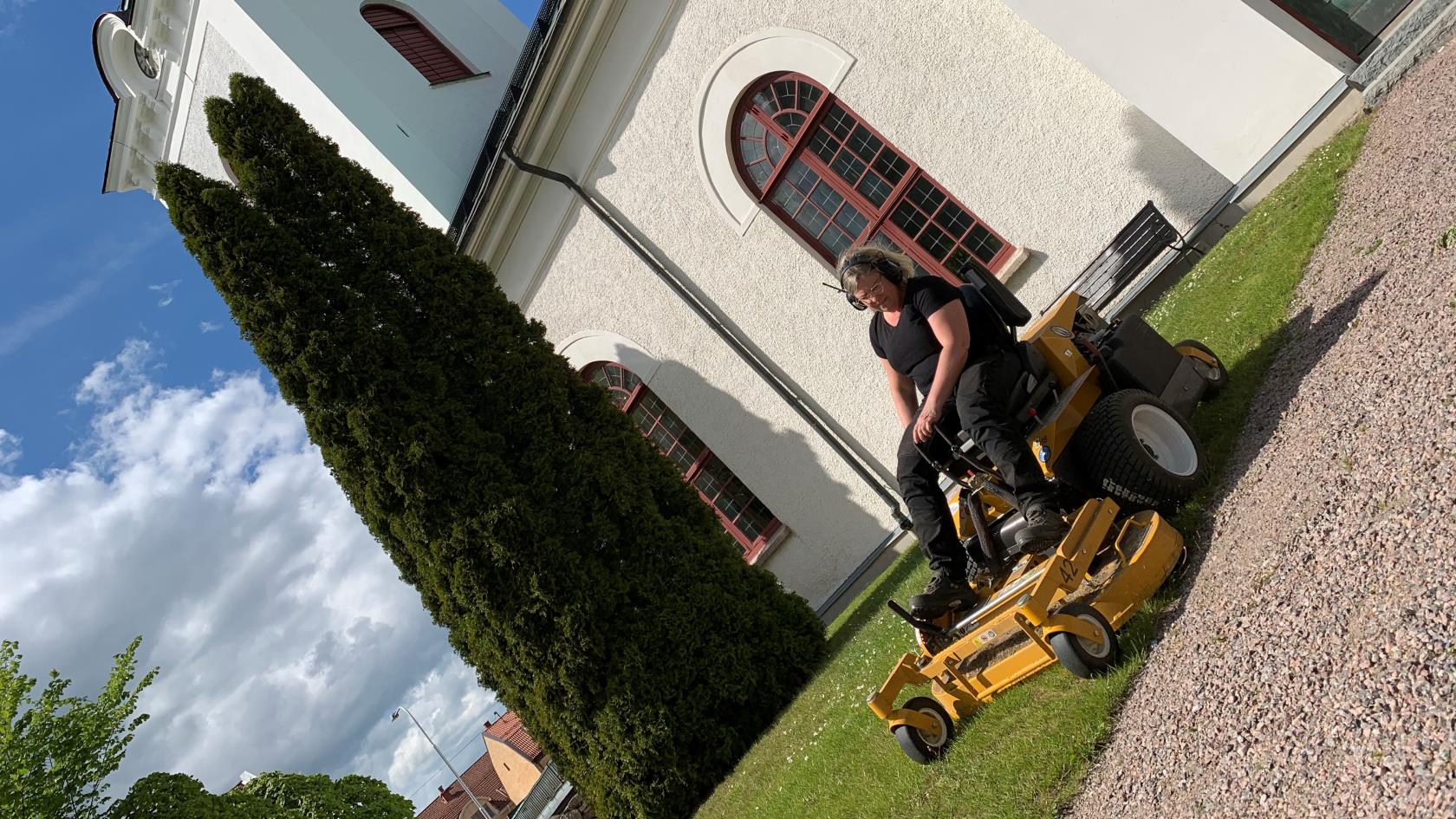 En snedtagen bild på en vaktmästare som kör gräsklippare på grästmattan framför en kyrka, bakom syns kyrkan med torn, dörr och fönster.