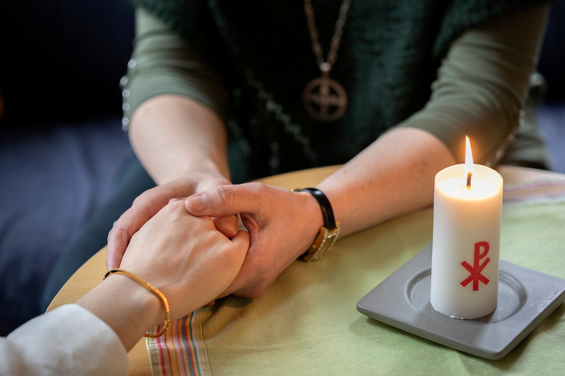 En diakon håller en annans hand i sina händer. Ett stearinljus med kristusmonogram brinner på bordet mellan dem.