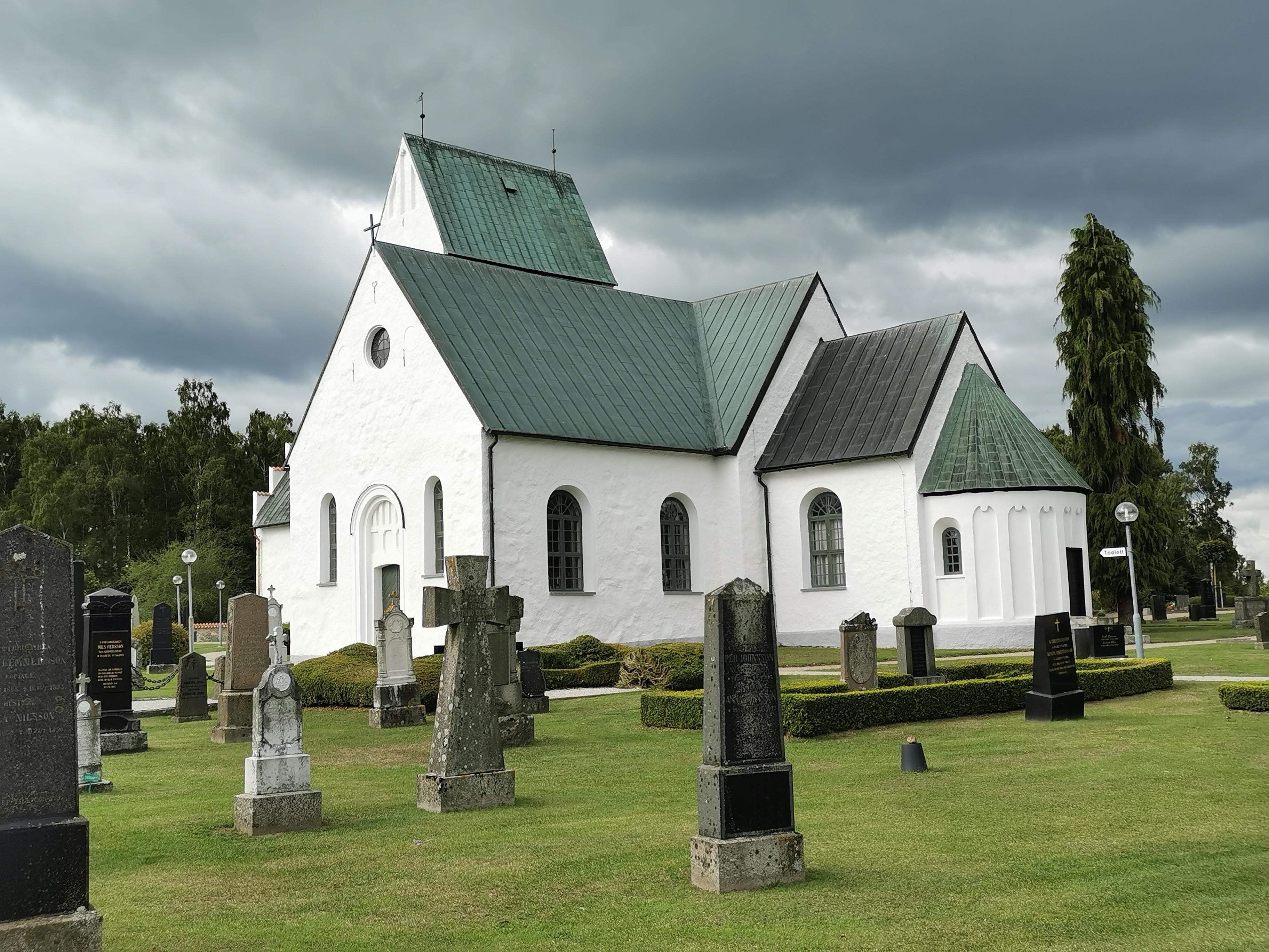 Köpinge kyrka med gröna träd i bakgrunden och svarta moln på himmelen.