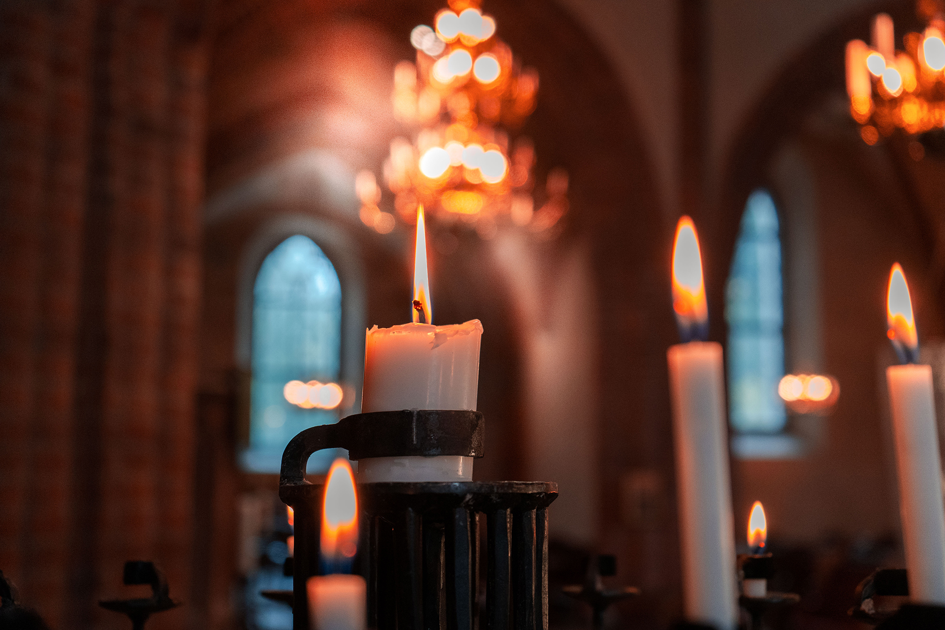 Flera tända ljus i en ljusbärare. I bakgrunden skymtar kyrkorummet och dess välvda fönster som också släpper in lite ljus. Ovanför ljusen glimmar ljussken från en av kyrkans gyllene takkronor.