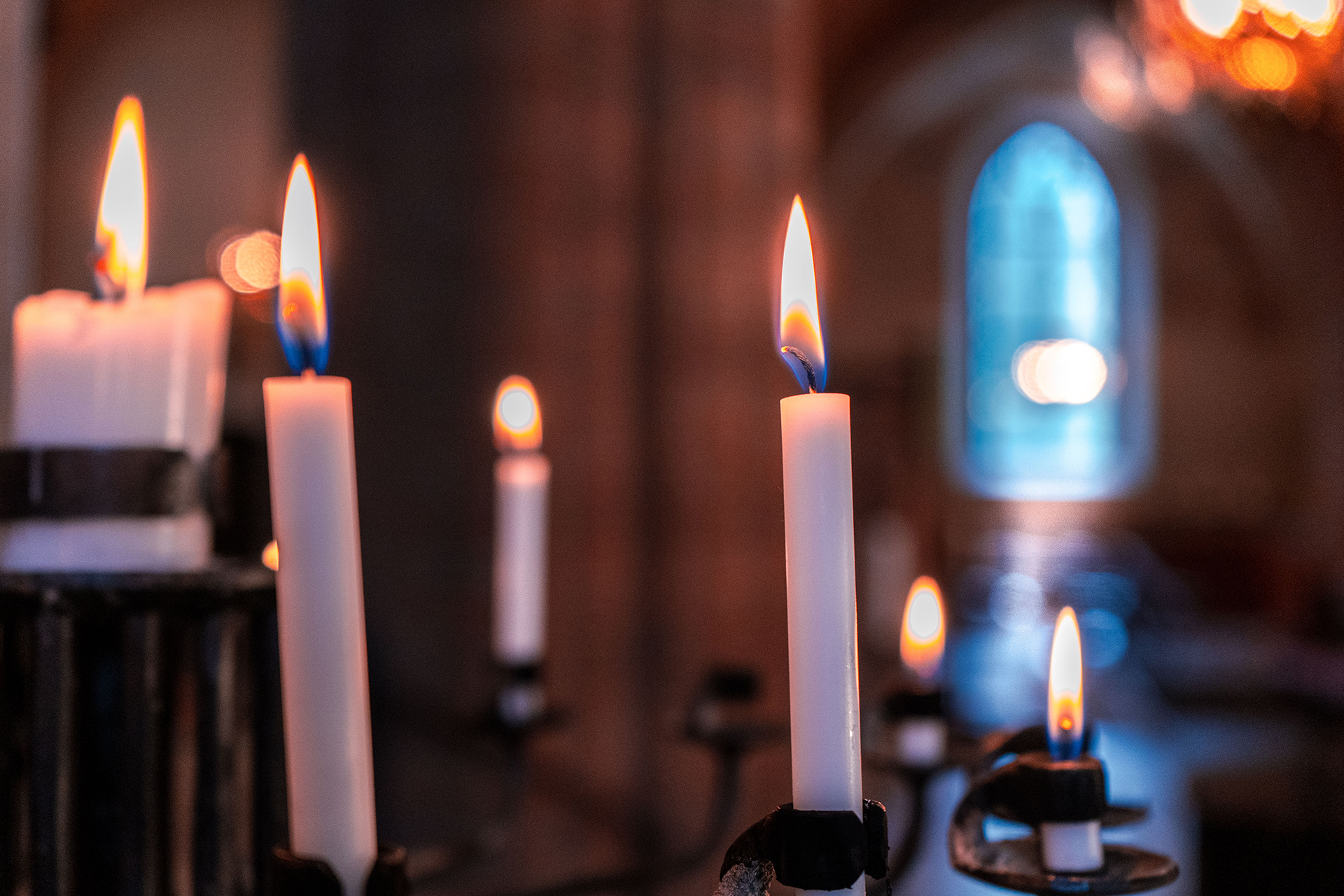 Flera tända ljus som står och glimmar i en ljusbärare. I bakgrunden skymtar kyrkorummet med dess välvda fönster som släpper in dagsljus.