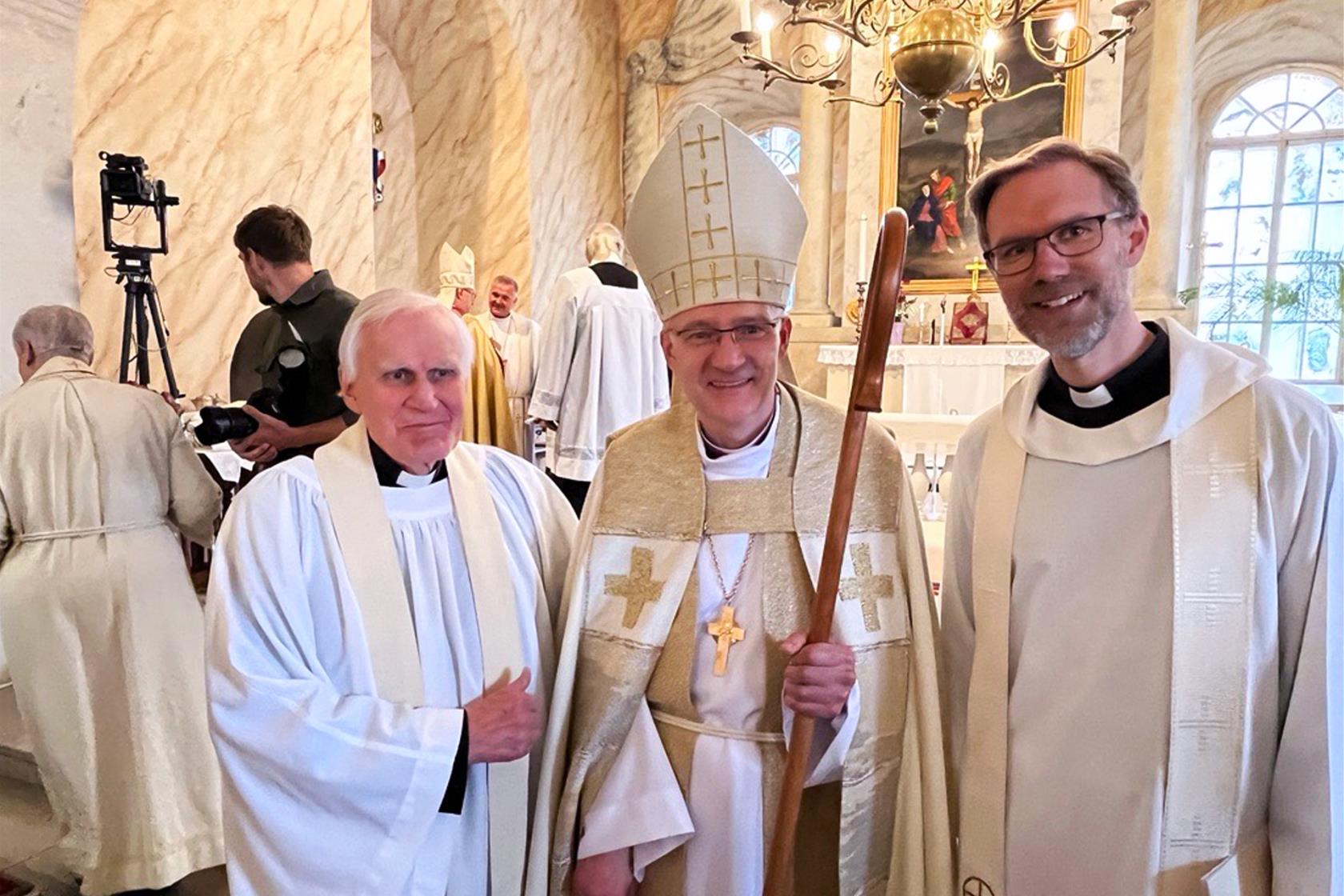 Tre vitklädda präster står framför ett altare och ler mot kameran: förre kyrkoherden i Gamla Uppsala församling, Per Anders Sölvin, nyvigda biskopen Anti Toplaan och nuvarande församlingsherden i Gamla Uppsala Andreas Holm.