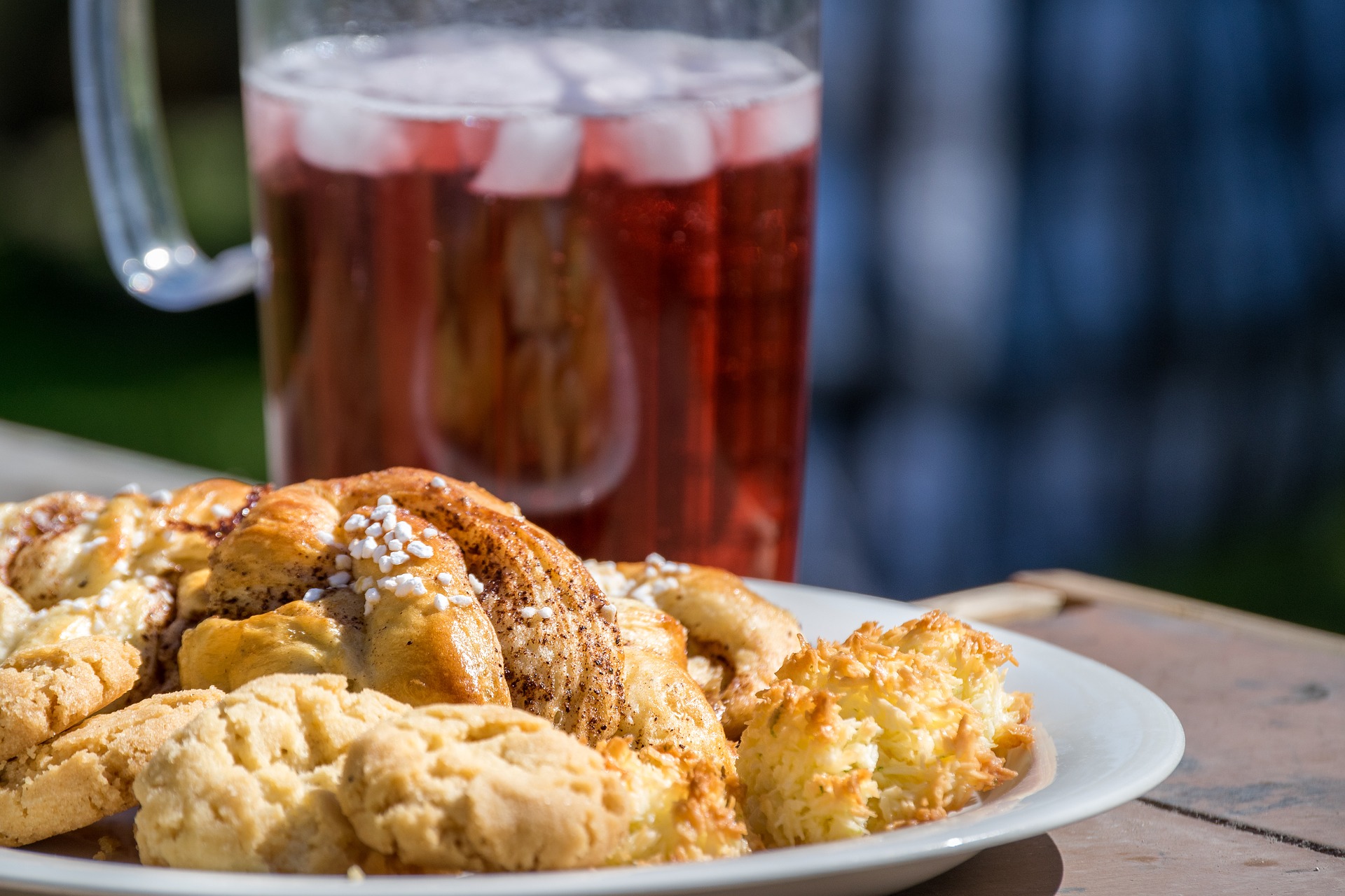 En fat med bullar och kakor står på ett bord utomhus och syns till vänster i bild. I bakgrunden syns en kanna med röd saft och grönt gräs.