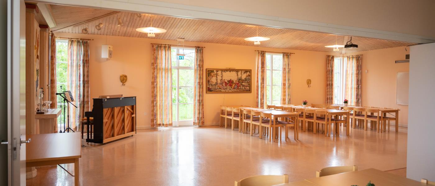 Vy över samlingssalen i Kimstads församlingshem. Till vänster står ett piano och till höger står långbord med stolar uppställda.