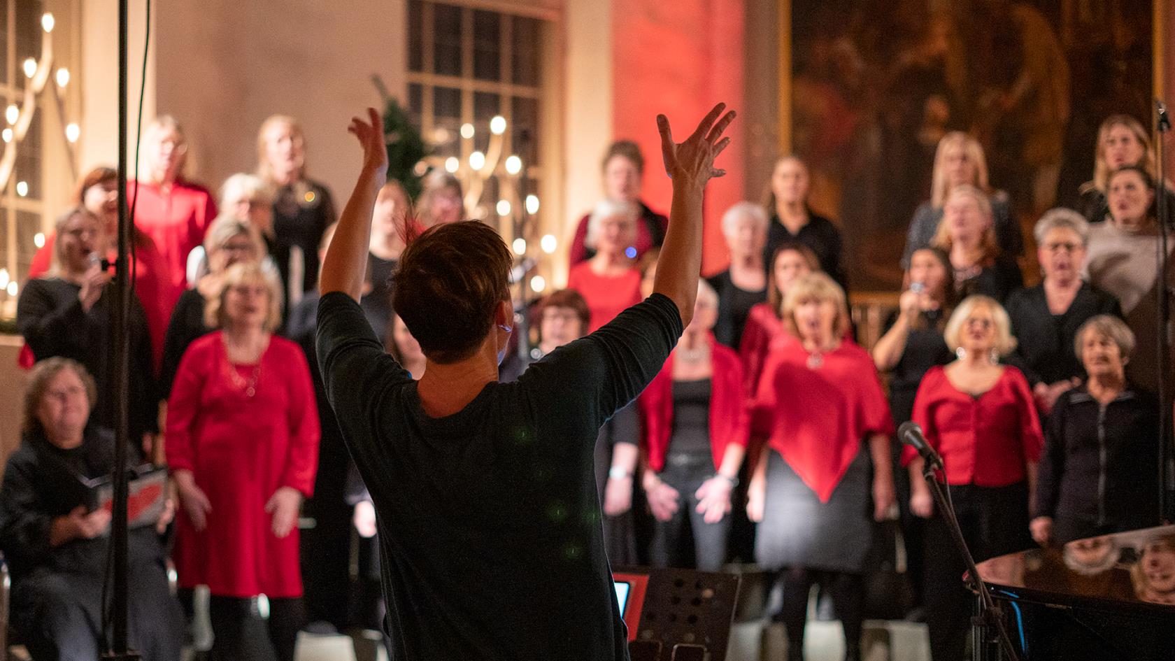 Körledare Malin Ekstrand syns bakifrån i skärpa, hon dirigerar kören. I oskärpa ser man en del av den stora kören Åby gospel. De är klädda i rött och svart. 