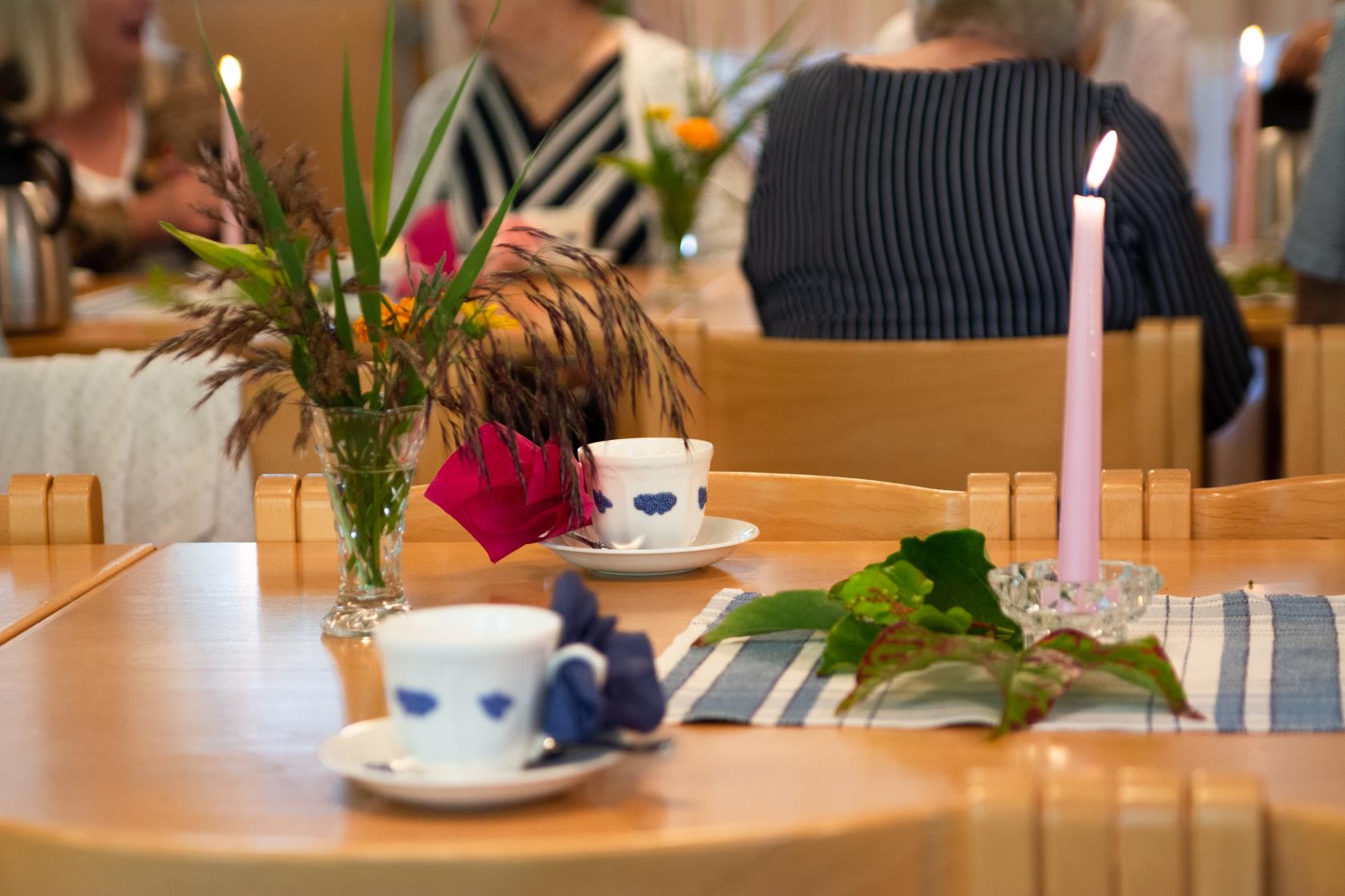 Ett uppdukat bord syns i bild. På bordet står brinnande ljusrosa ljus, vaser med gröna växter och blåvita kaffekoppar med servetter instuckna i örat. I bakgrunden syns kvinnor som samtalar och dricker kaffe.