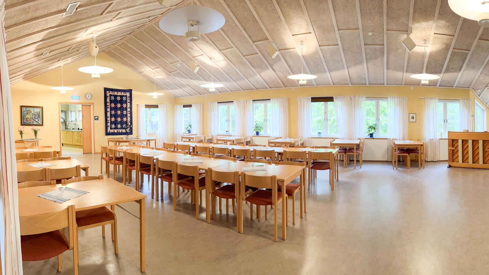 Panoramabild över församlingssalen. Till vänster står långbord och bord uppradade, till vänster är golvytan öppen. Bakom står ett piano och en soffgrupp utställt.