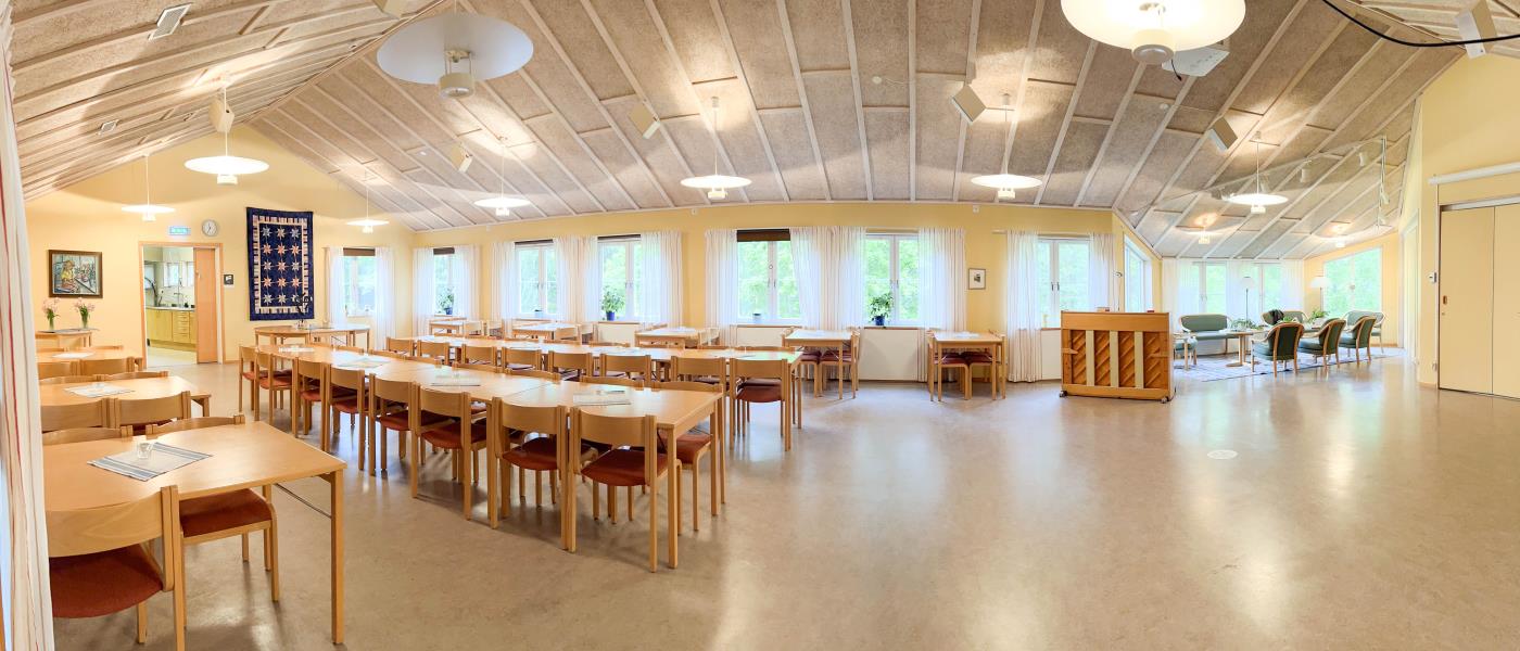 Panoramabild över församlingssalen. Till vänster står långbord och bord uppradade, till vänster är golvytan öppen. Bakom står ett piano och en soffgrupp utställt.