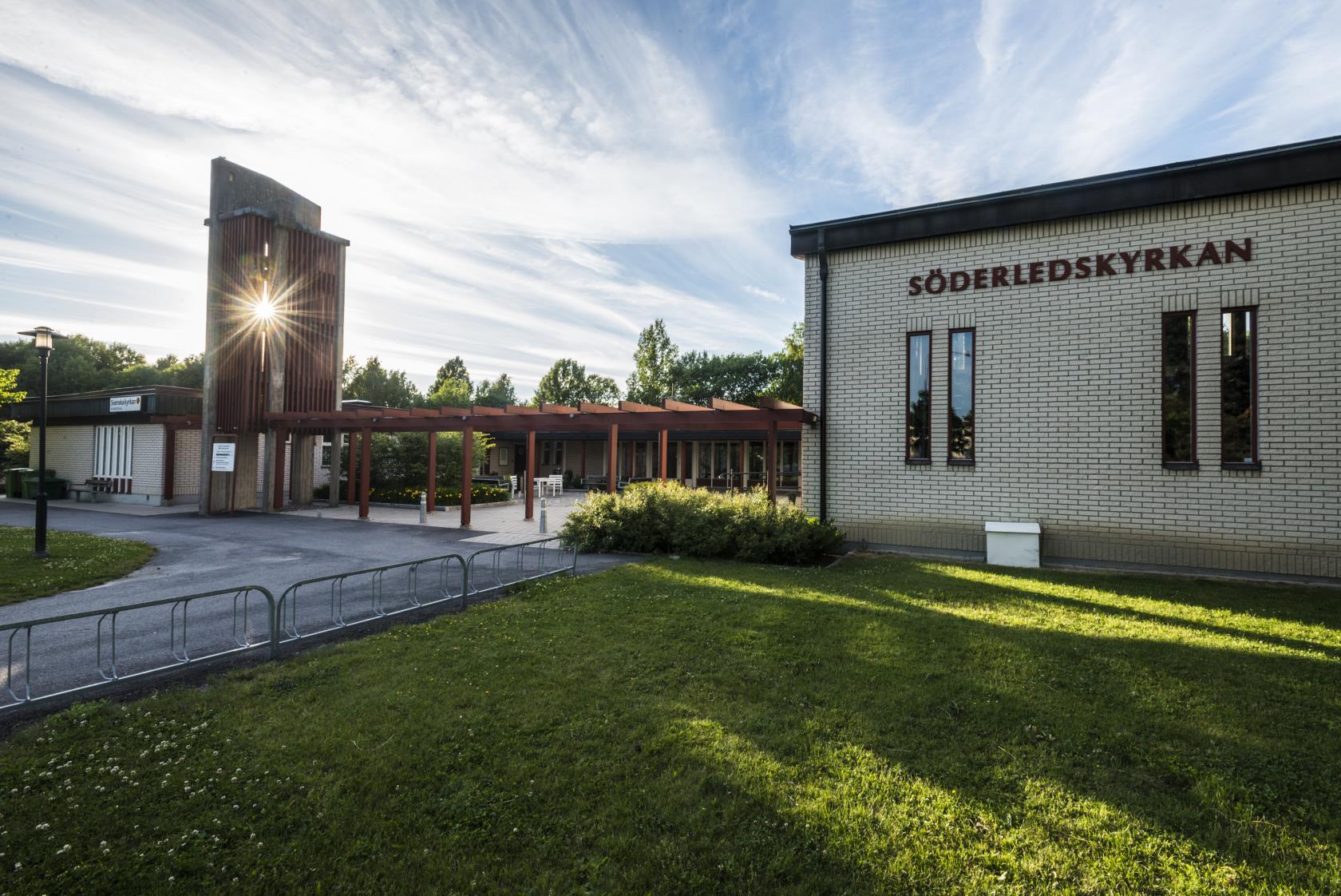 Söderledskyrkan en sommardag. Till höger i bild syns kyrkbyggnaden med texten Söderledskyrkan och till vänster syns den rödmålade klockstapeln. På himlen syns slöjmoln och bakom klockstapeln skiner solen igenom.