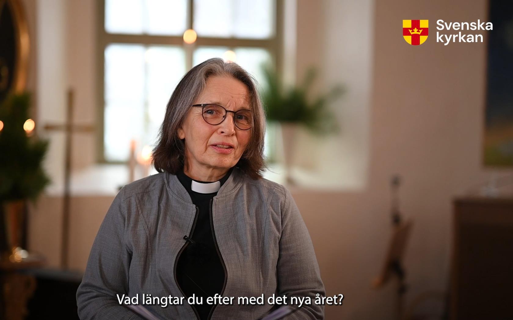 Präst Eva Strannelid står och talar med kyrkans interiör i bakgrunden. Ett fönster, ett kors, ljus i oskärpa.