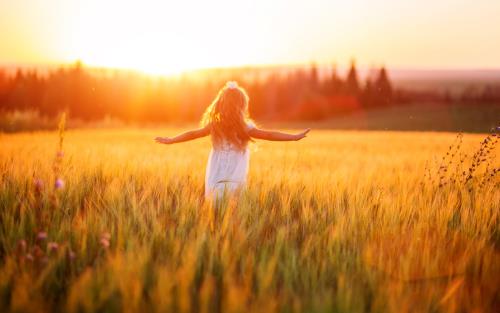En flicka springer med utsträckta armar över ett gyllene sädesfält i solnedgång.
