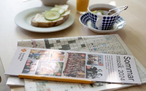 Ett uppdukat frukostbord. På bordet ligger en tidning med korsord och en sommarmusikfolder.