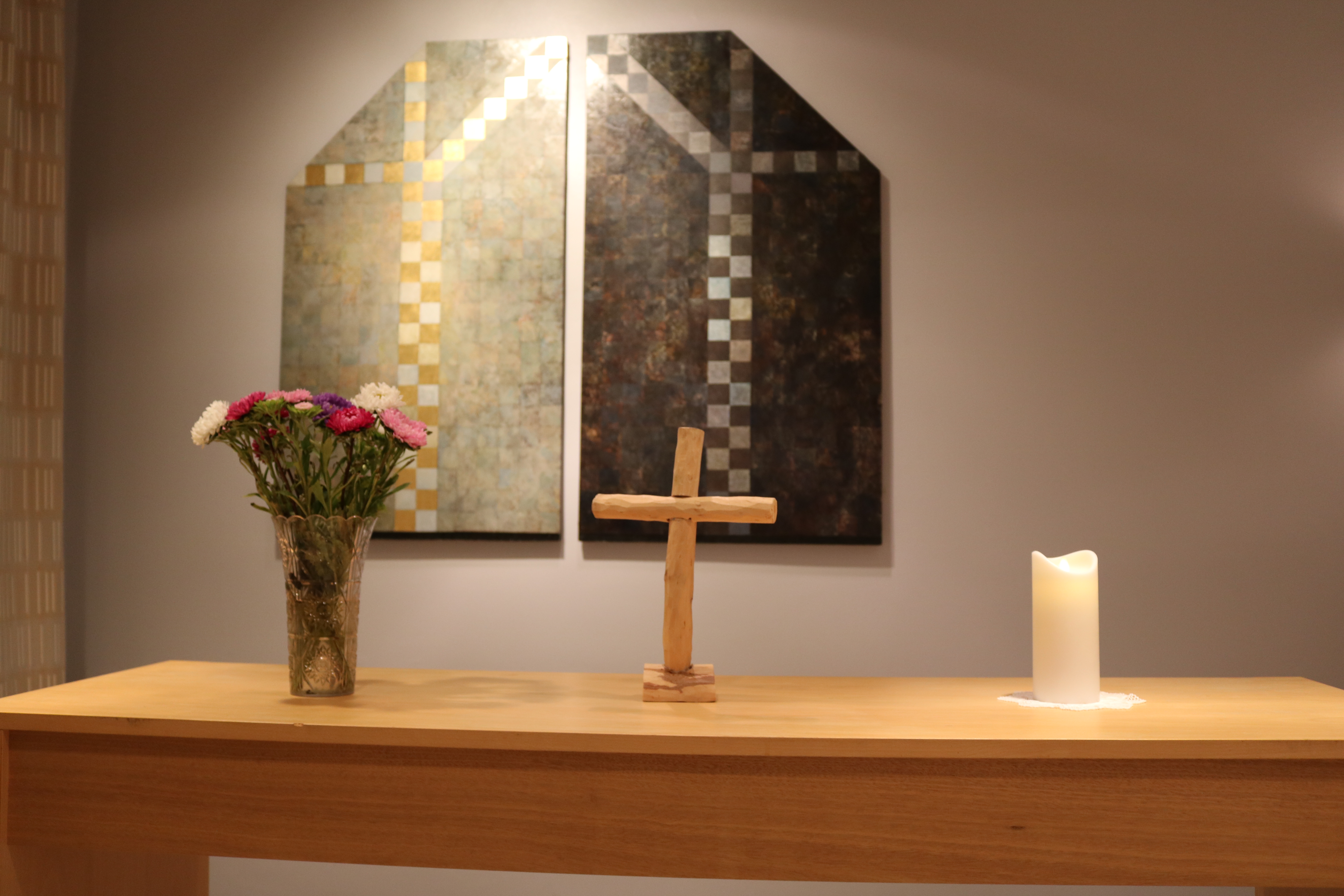 Kyrkrummet i Mölndals sjukhus. På väggen hänger en tvådelad tavla med mosaik som ett fönster. På altaret framför står en vas med blommor, ett träkors och ett tänt ljus. 