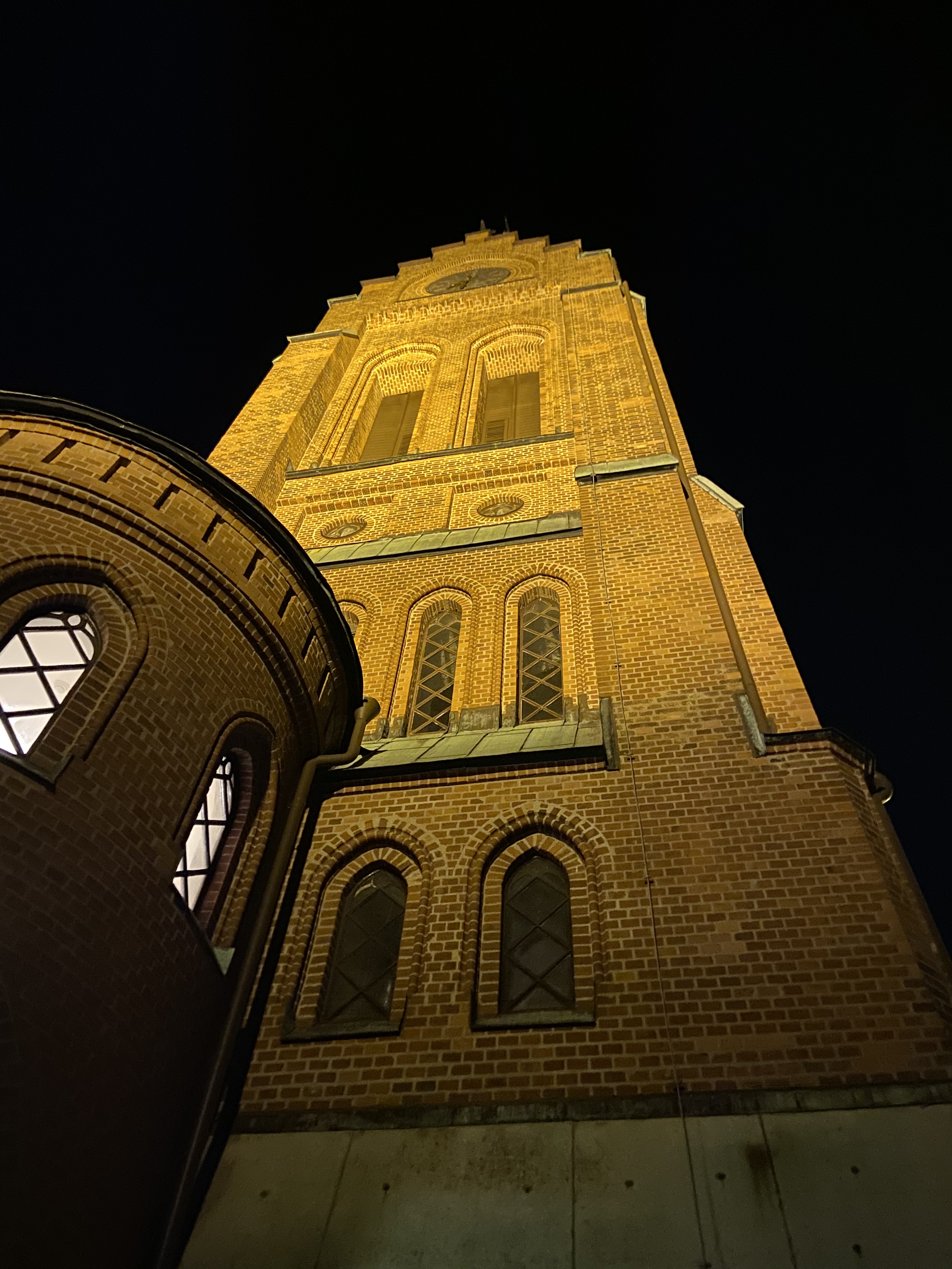 Fässbergs kyrktorn fotograferat nerifrån på kvällen. Mörk bakgrund, tornet är upplyst.