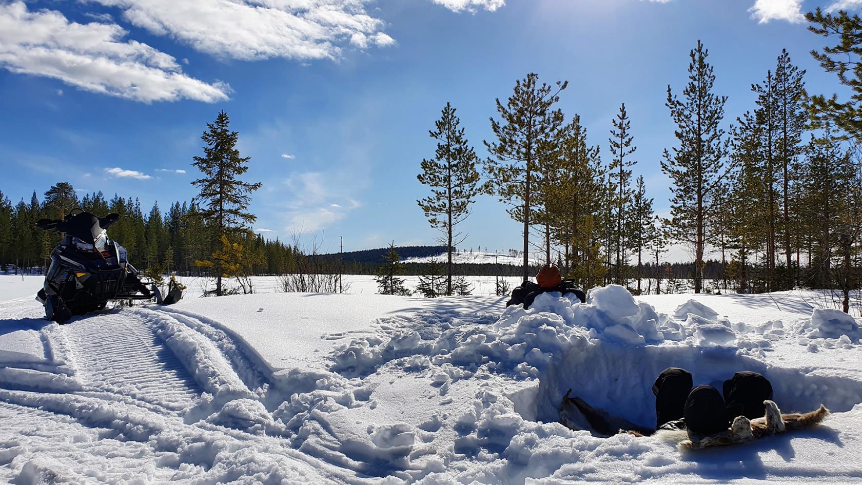 Två personer sitter i varsin grop i en snödriva ute i skogen och solar sig.  Intill står en skoter parkerad. 