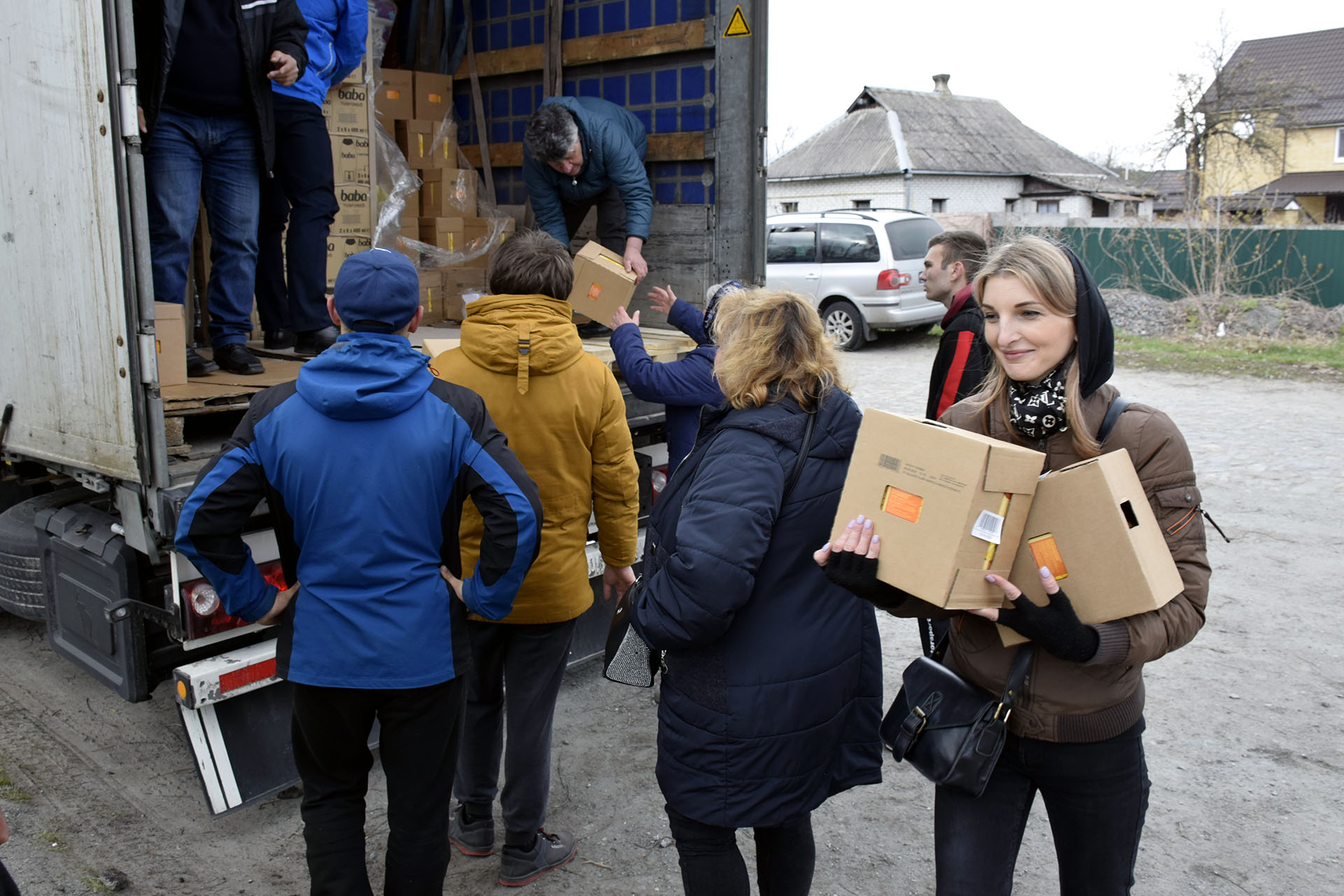 En kvinna går förbi kameran med paket i famnen. Bakom henne står det ett antal personer och väntar på att få sina paket som lastas ut av tre personer som står inne i lastbilen. 