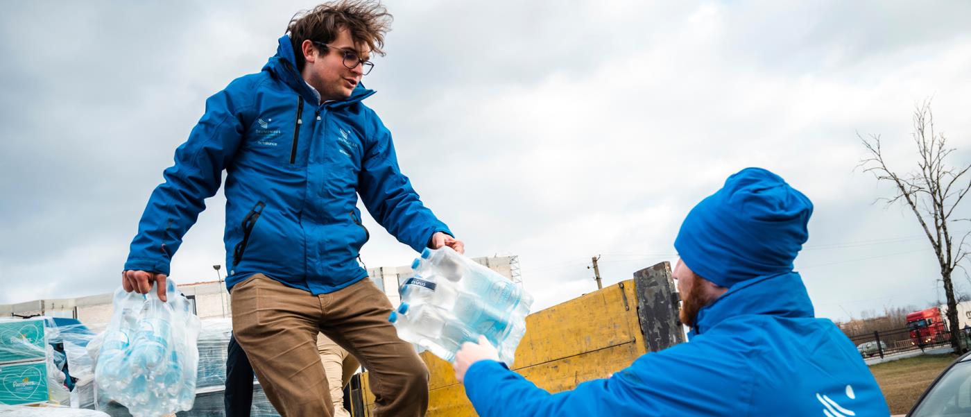 Vattenflaskor lyfts av från flaket av en last bil. Två män i blåa organisationsjackor.