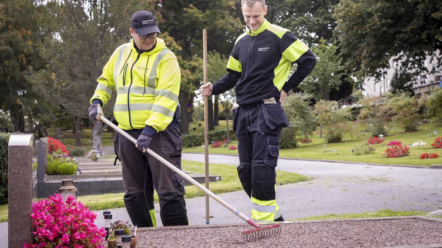 Två män i varselkläder och med krattor vid en gravplats med gravsten och rosa blommor.