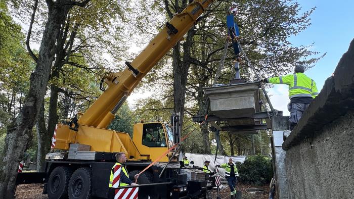 Lyftkran lyfter stenportal på plats uppe på pelare på kyrkogård med träd runtomkring.