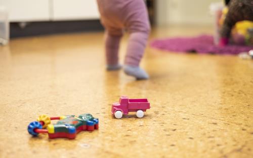 Leksaker på ett golv. I bakgrunden två ben på litet barn klätt i rosa leggings. 