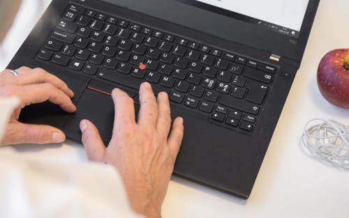 En kvinna skriver på en bärbar dator. Bredvid datorn ligger ett äpple, vita hörlurar och två godispaket. Fotot är taget över kvinnans axel.