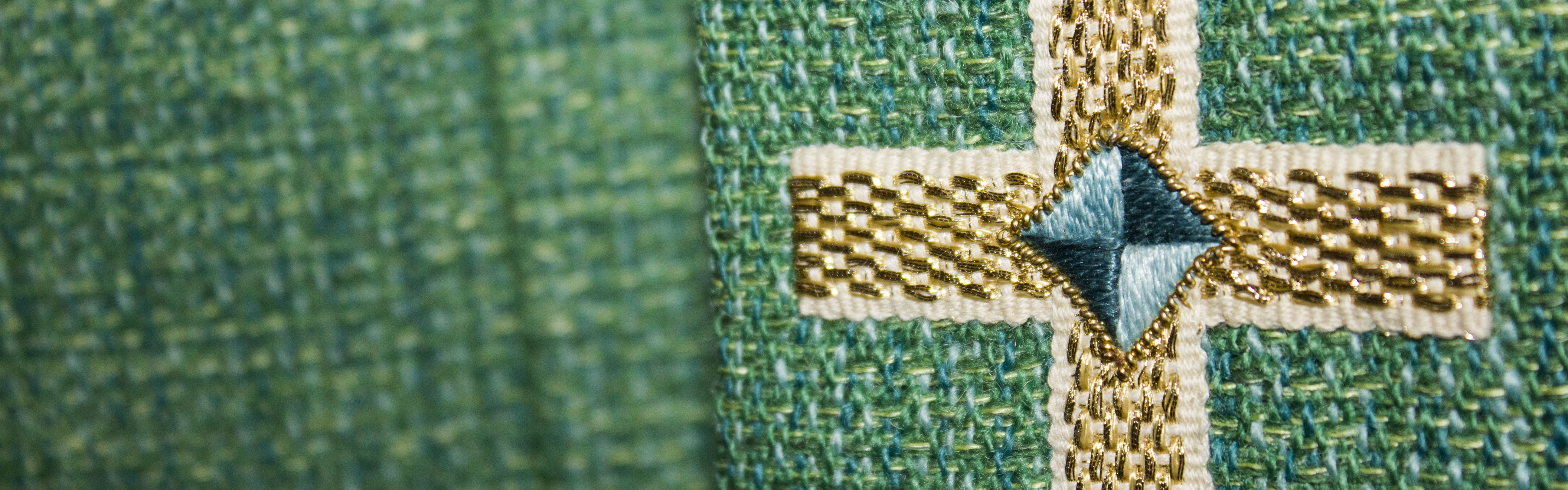 Ett broderat kors i guld, vitt och blått på grönt tyg.