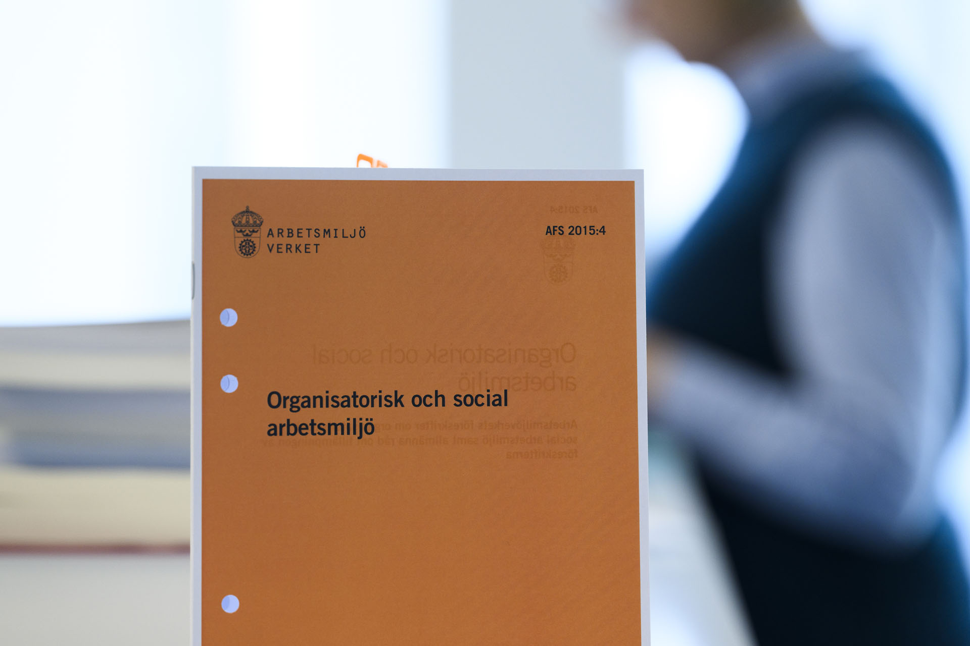 Folder om organisatorisk och social arbetsmiljö i förgrund, person i bakgrund. 
