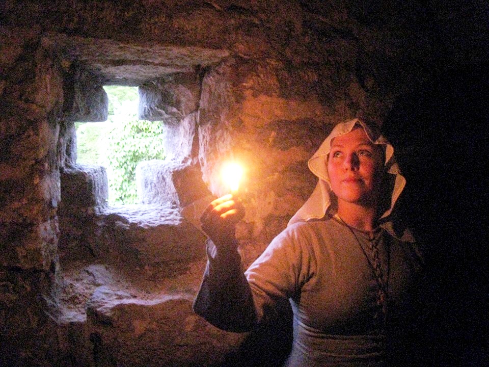 Kvinna i en krypta med litet ljus i handen. Korsformad fönsterglugg släpper in lite ljus