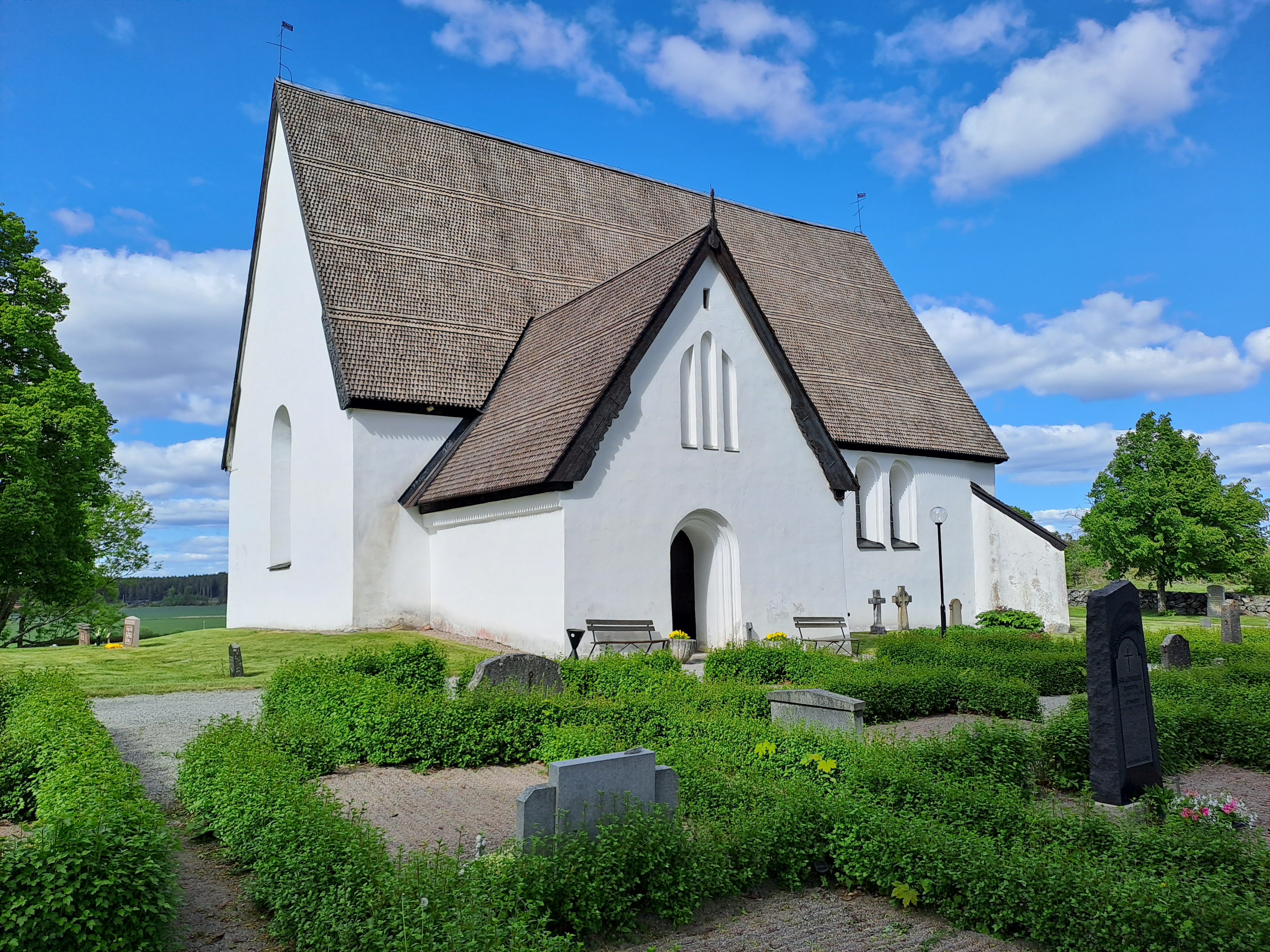 En vit medeltida stenkyrka med svart tak. Himlen är klarblå, gräset och växterna runt kyrkan är gröna.