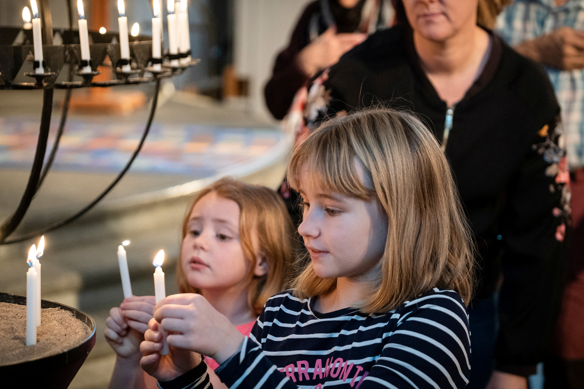 Två barn som ser ut att vara fem och sju år gamla tänder ljus vid en ljusbärare i ett kyrkorum.