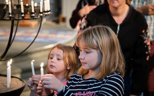 Två barn som ser ut att vara fem och sju år gamla tänder ljus vid en ljusbärare i ett kyrkorum.