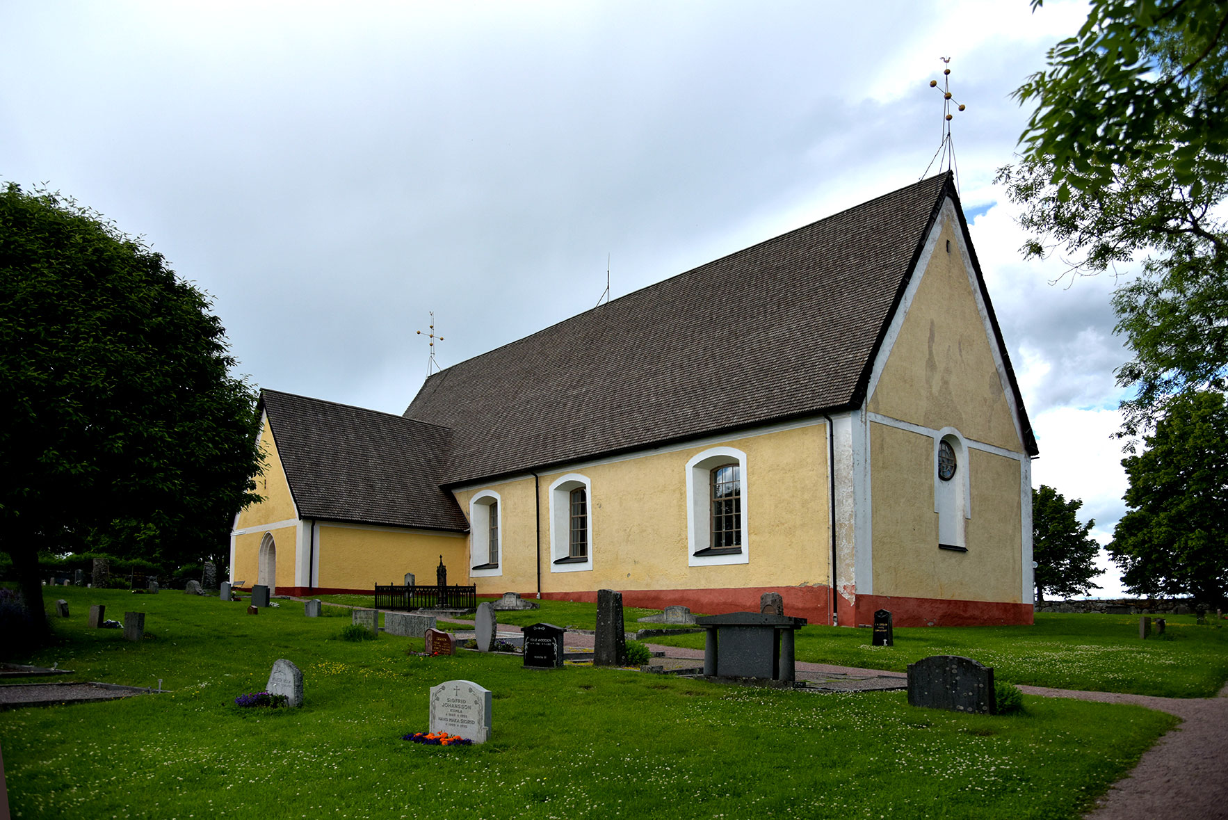 En gul kyrka utan torn. I förgrunden gräsmatta med några gravar. 