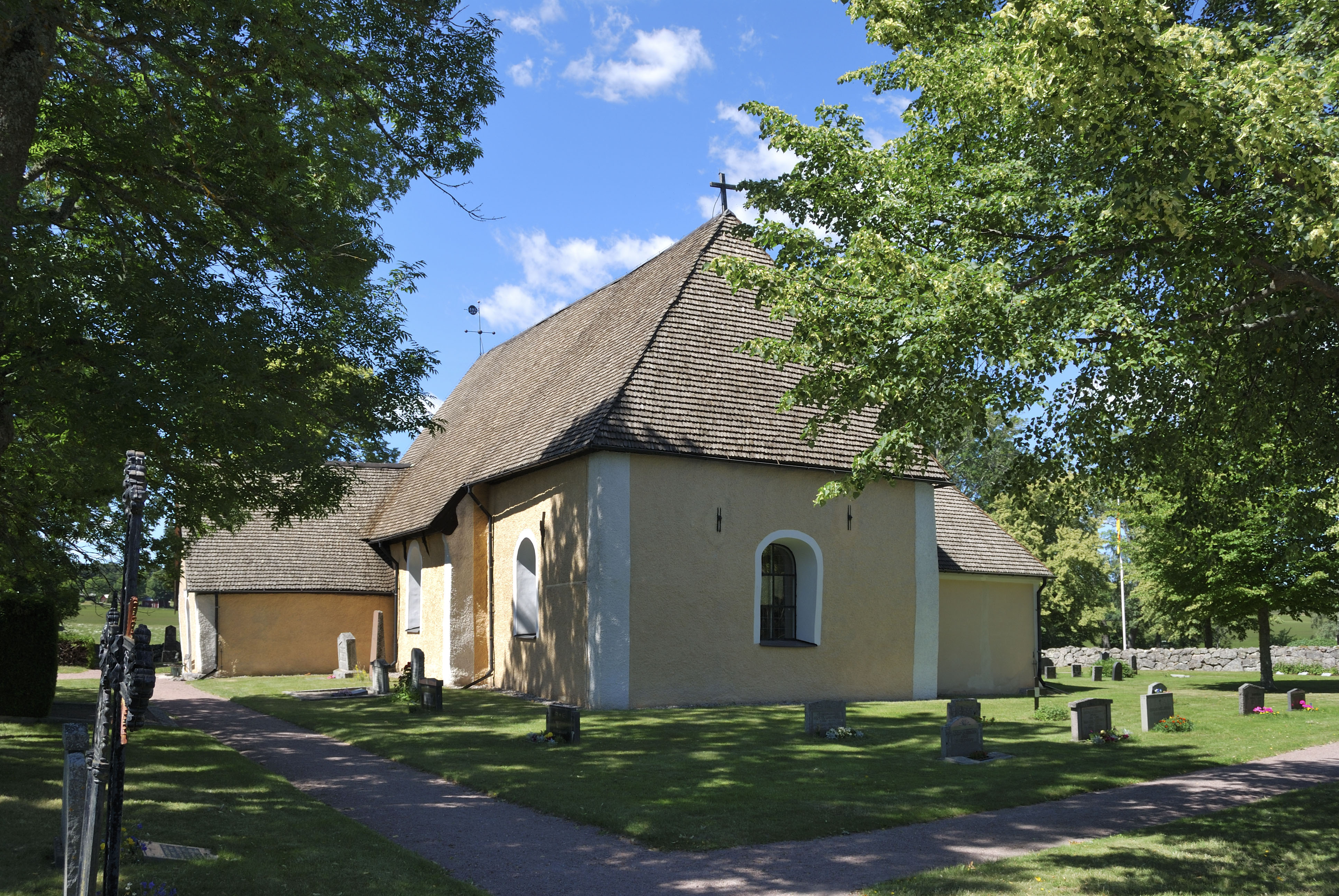 En liten gul stenkyrka, omgiven av gröna träd.