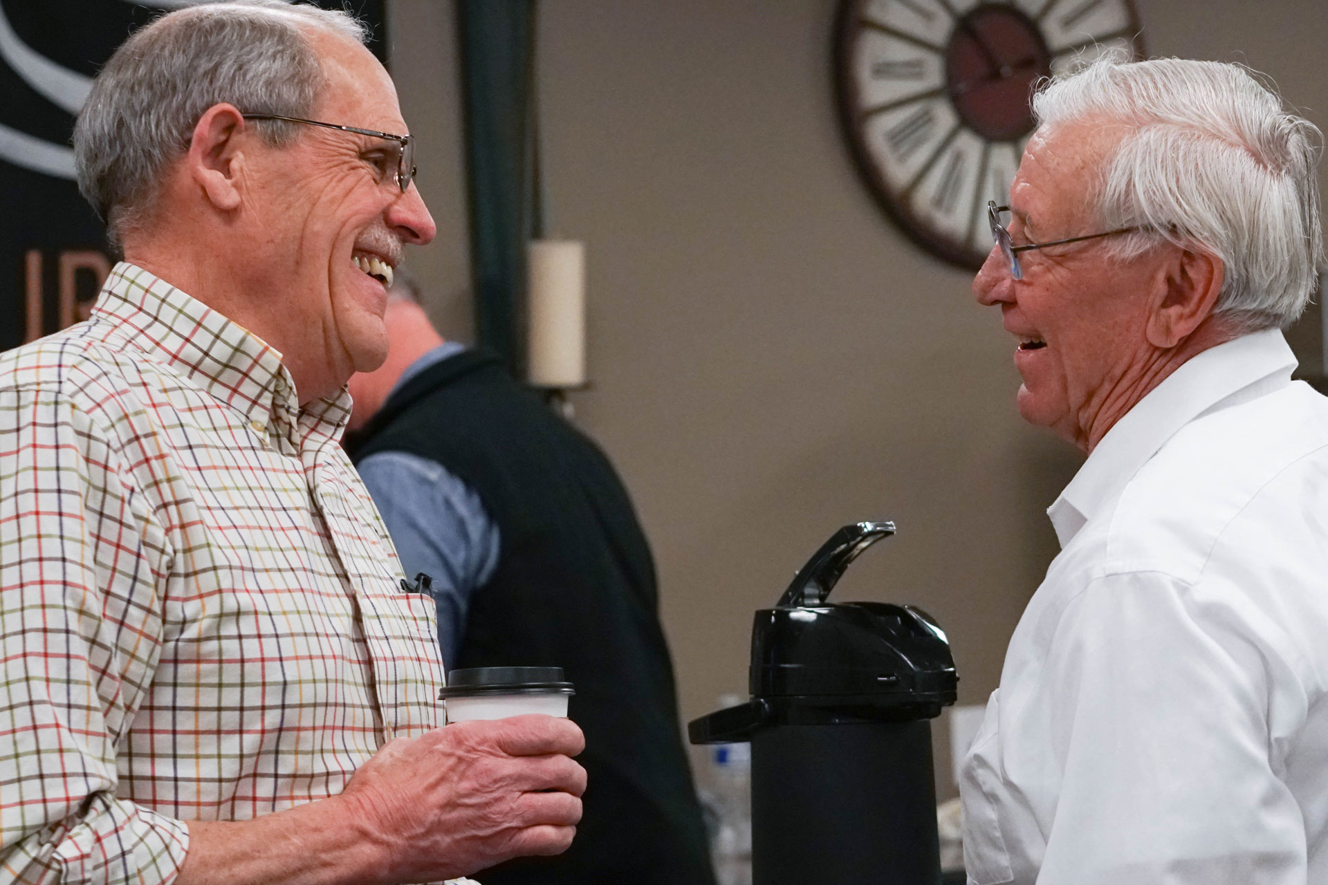 Två äldre män samtal och ser glada ut. Den ena håller en pappmugg. 