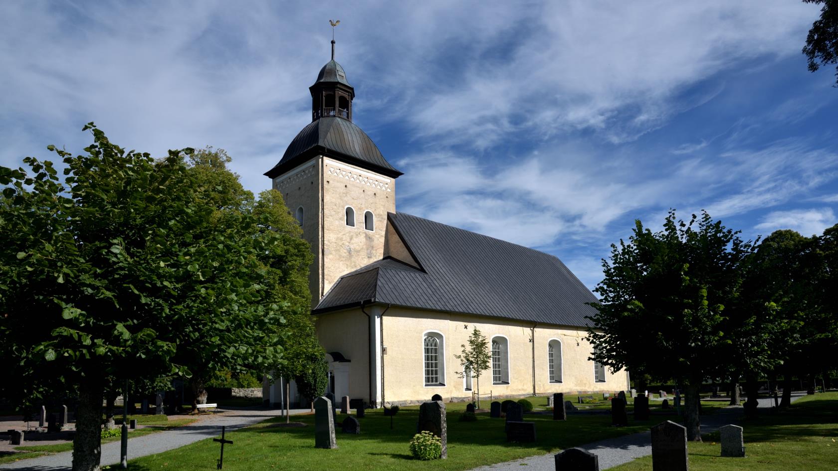 En gulaktig kyrka med ett torn. Gröna lövträd står på kyrkogården runt kyrkan. Himlen är blå.