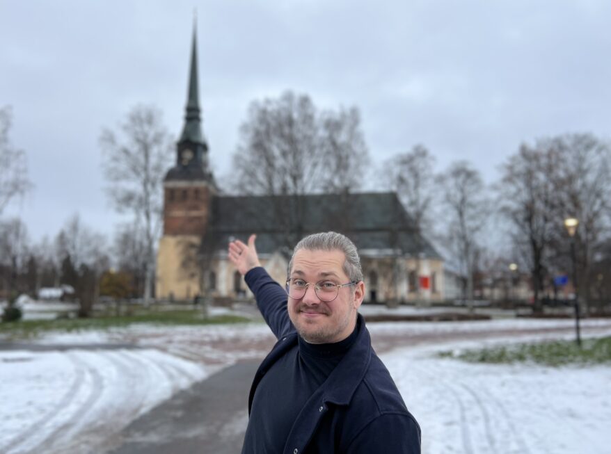 man står framför mora kyrka och ler medans han pekar med hela handen mot kyrkan 