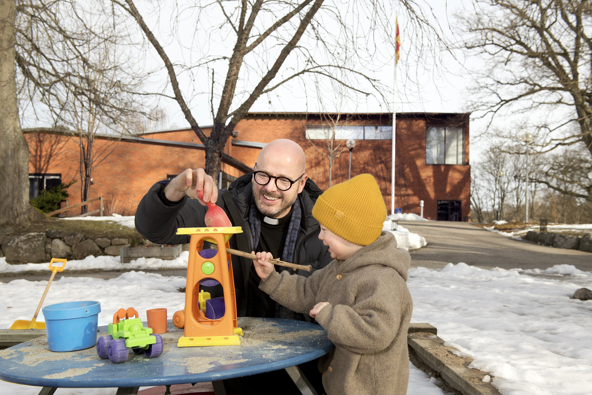 Bilden visar Adolfsbergs kyrka i bakgrunden. I förgrunden syns en präst och ett barn som leker vid lekplatsen.