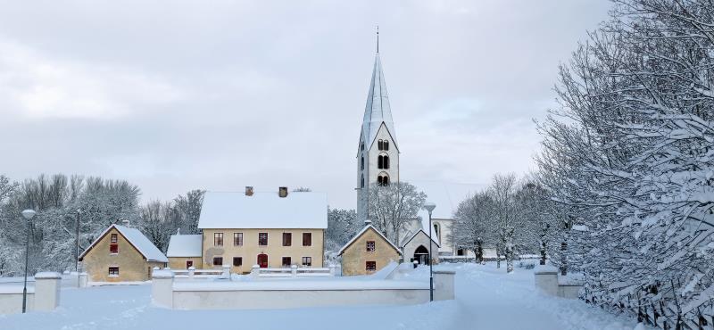 Othem prästgård och kyrka från söder. Prästgården är ljusgul. Kyrkans torn sticker upp högt. Snön ligger mjukt på marken. 