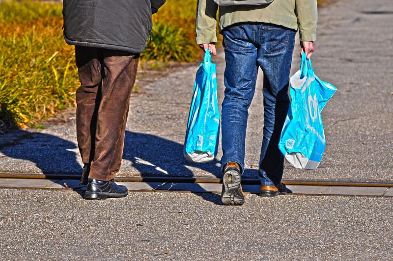 Två personer går på en väg, med ryggarna mot oss. Den ene bär matkassar.